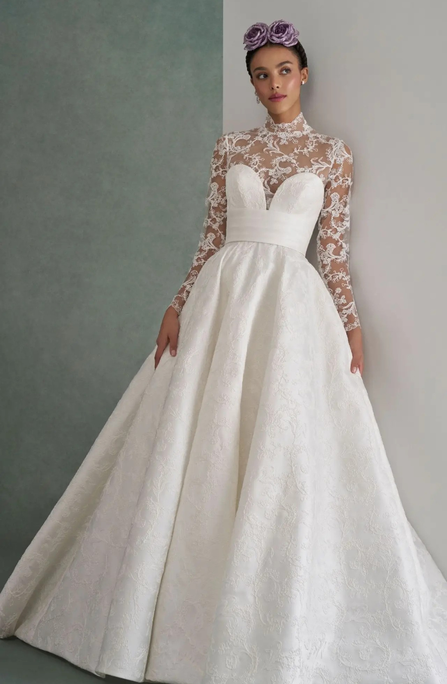 Model is wearing a ball gown with a long sleeve lace underlay with a high neckline, a fitted bodice, and plunging neckline by mark ingram