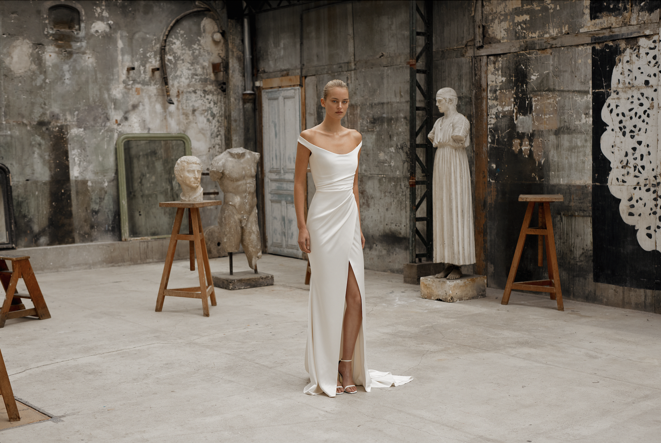 model is wearing a sheath off the shoulder wedding dress with a high slit and minimalist fabric