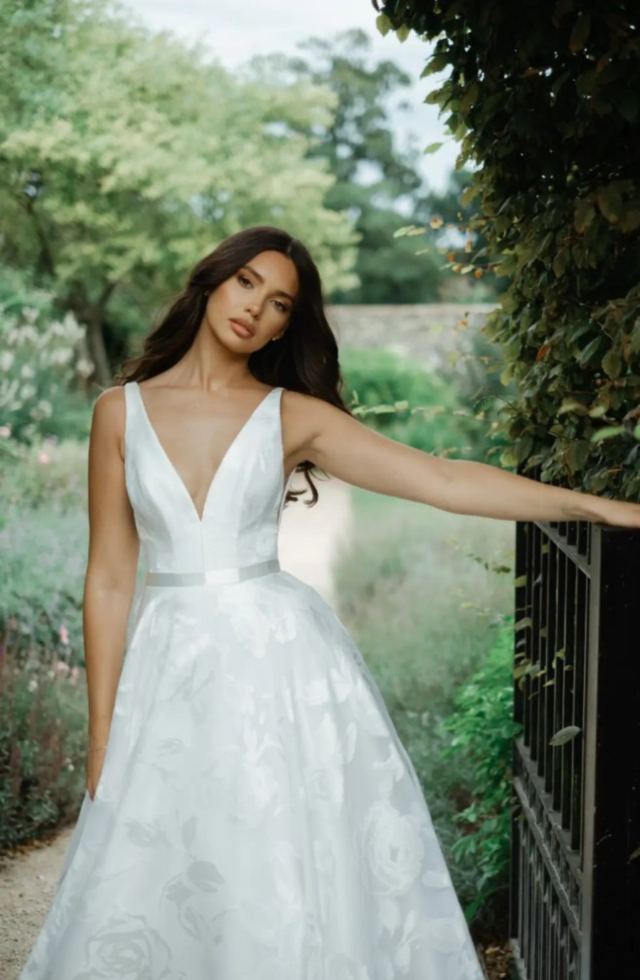Model is wearing a sleeveless a-line bridal gown with a floral embroidered skirt, plunging neckline, and satin belt by suzanne neville